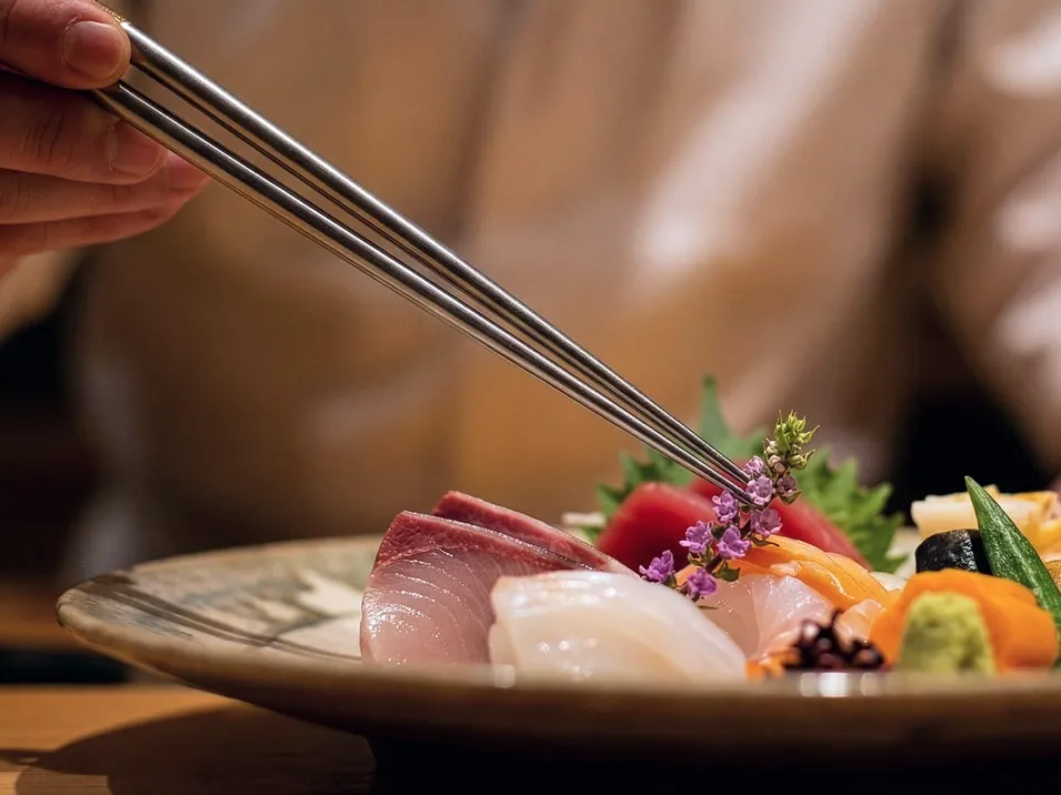 皿に盛られた鮮魚のお造りと、その上に銀の箸で丁寧に紫色の花穂（穂紫蘇）を添える料理人の手元。