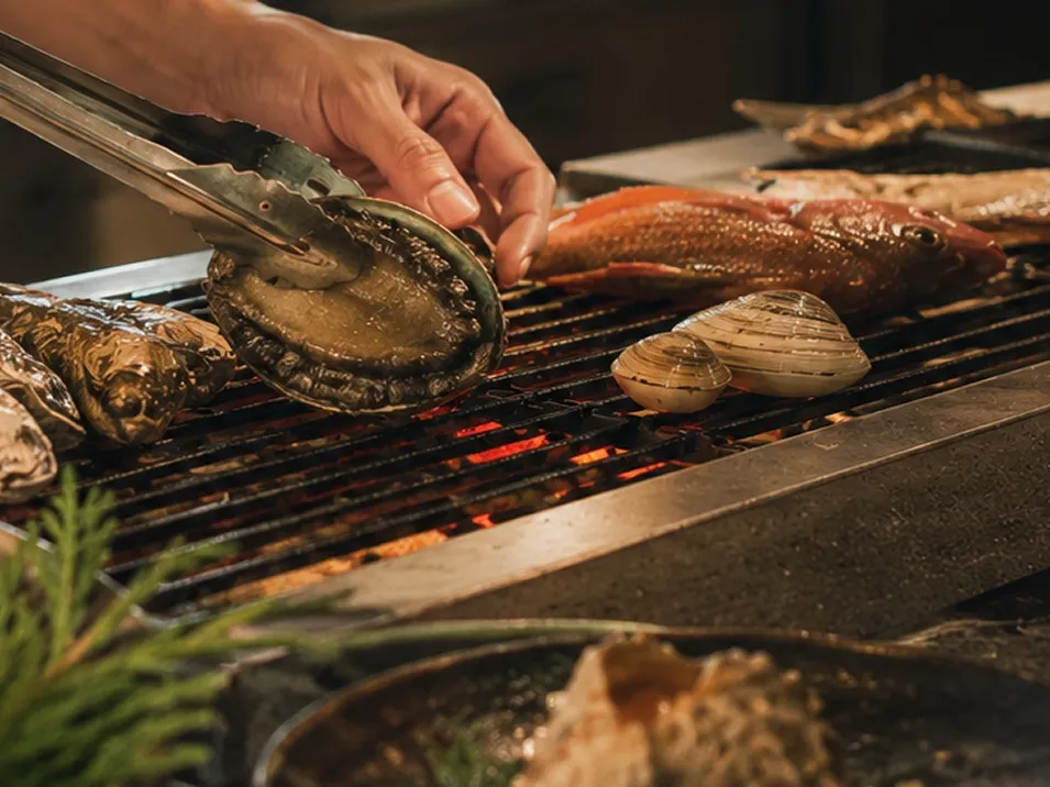 赤赤と起きる炭火の網の上で、トングを使いアワビやハマグリ、魚をじっくりと焼き上げる調理の手元。