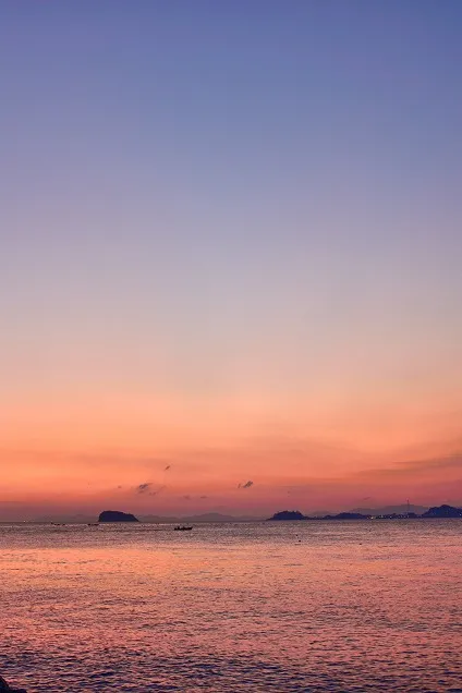 夕暮れ時のグラデーションに染まる空と水面、水平線にたたずむ島影と小さな船の情景。